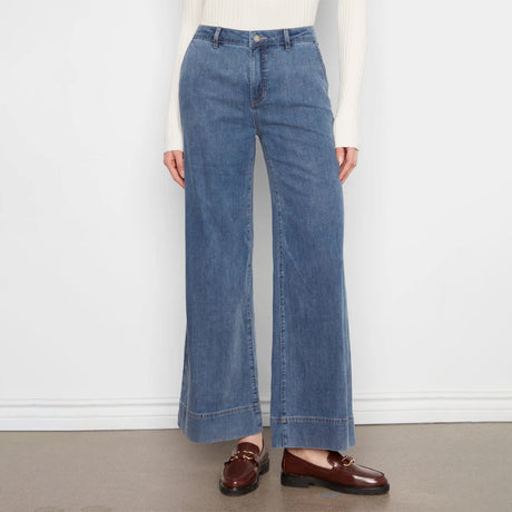 A model wearing a pair of wide-leg blue denim jeans with a rolled hem, styled with a cream ribbed long-sleeve top and brown leather loafers against a white wall and gray floor.