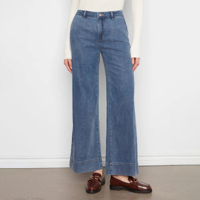 A model wearing a pair of wide-leg blue denim jeans with a rolled hem, styled with a cream ribbed long-sleeve top and brown leather loafers against a white wall and gray floor.
