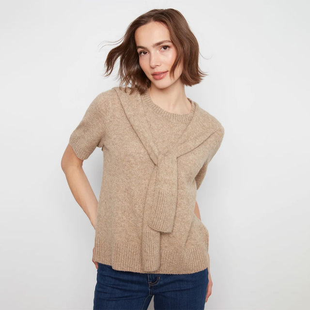 A woman wearing a beige short-sleeve knit sweater with a draped front detail, paired with dark blue jeans, against a plain light gray background.