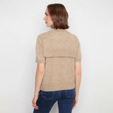 A woman shown from the back wearing a beige short-sleeve knit top with a ribbed band across the upper back and blue jeans, against a plain light gray studio background.