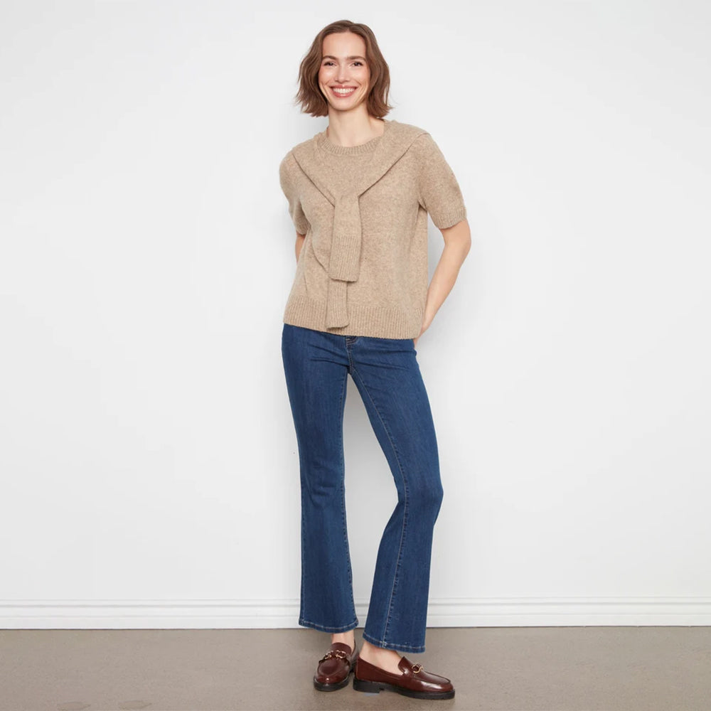 A woman modeling a beige knit short-sleeve top with a draped front detail, paired with dark blue flared jeans and brown leather loafers, standing against a plain white wall.