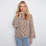 A woman stands against a white wall wearing a textured, multicolor knit sweater with beige, brown, and gray tones, paired with light blue jeans.
