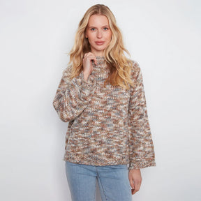 A woman stands against a white wall wearing a textured, multicolor knit sweater with beige, brown, and gray tones, paired with light blue jeans.