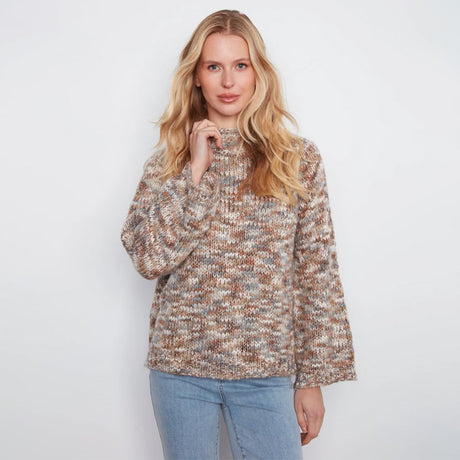 A woman stands against a white wall wearing a textured, multicolor knit sweater with beige, brown, and gray tones, paired with light blue jeans.