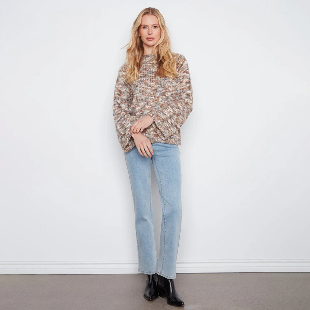 A woman stands against a white wall wearing a textured, multicolor knit sweater with beige, brown, and gray tones, paired with light blue jeans and black ankle boots.