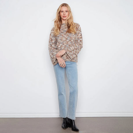 A woman stands against a white wall wearing a textured, multicolor knit sweater with beige, brown, and gray tones, paired with light blue jeans and black ankle boots.