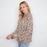 A woman stands against a white wall wearing a textured, multicolor knit sweater with beige, brown, and gray tones, paired with light blue jeans.