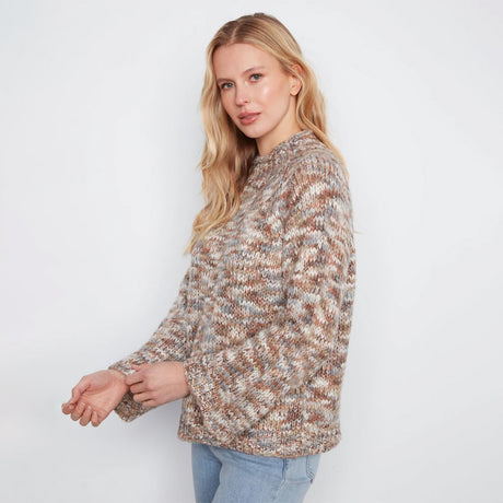 A woman stands against a white wall wearing a textured, multicolor knit sweater with beige, brown, and gray tones, paired with light blue jeans.