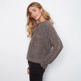 A woman modeling a chunky knit crewneck sweater in a heathered brown and gray color, paired with black pants, against a plain white studio background.