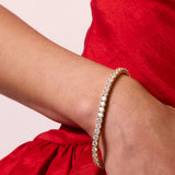 A close-up of a gold bangle bracelet with a line of clear pavé stones worn on a person's wrist, paired with a red dress against a light beige background.