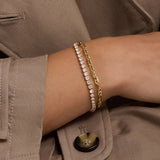 A close-up of a person's wrist wearing two delicate bracelets: one gold chain bracelet and one gold tennis bracelet with clear stones, styled with a taupe blazer.