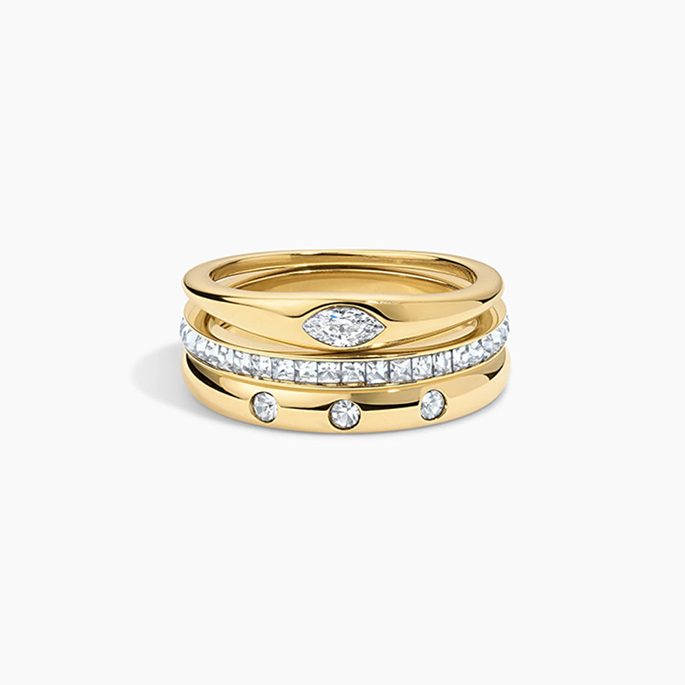 A stack of three gold rings on a light gray background. The top ring is plain gold, the middle ring is paved with clear stones, and the bottom ring is gold with three small clear stones spaced along it.