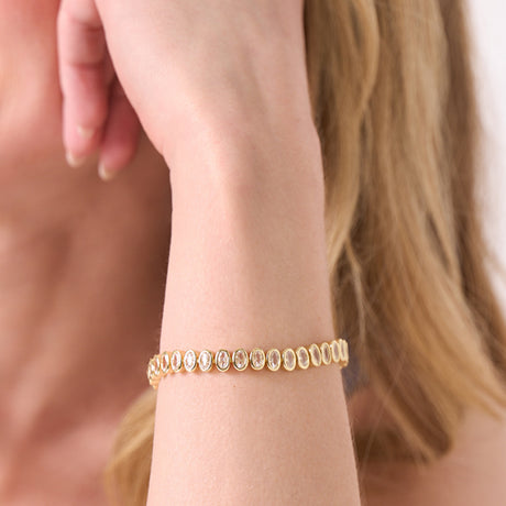 A close-up of a woman's wrist wearing a delicate gold bracelet with small oval-cut clear stones, shown against her skin and blonde hair.