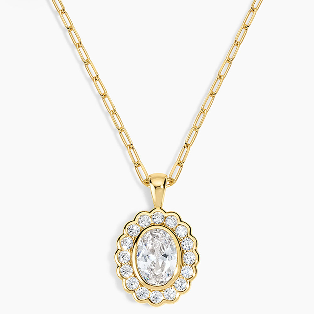 A gold pendant necklace featuring a scalloped oval frame with clear stones surrounding an oval center stone, suspended from a gold paperclip chain on a light gray background.