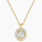 A gold pendant necklace featuring a scalloped oval frame with clear stones surrounding an oval center stone, suspended from a gold paperclip chain on a light gray background.