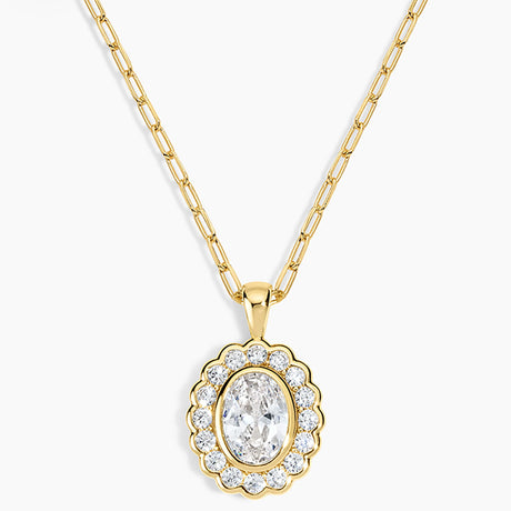 A gold pendant necklace featuring a scalloped oval frame with clear stones surrounding an oval center stone, suspended from a gold paperclip chain on a light gray background.