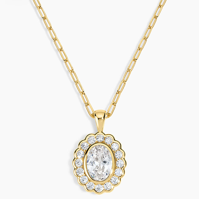 A gold pendant necklace featuring a scalloped oval frame with clear stones surrounding an oval center stone, suspended from a gold paperclip chain on a light gray background.