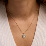 A close-up of a person's neck and chest wearing a delicate gold chain necklace with an oval pendant set with clear stones, paired with a cream-colored knit top.