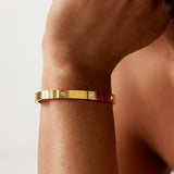 A close-up of a gold bangle bracelet with three small clear stones, worn on a person's wrist against a light neutral background.