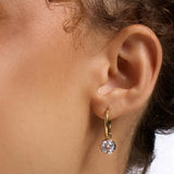 A close-up of a person's ear wearing a delicate gold huggie hoop earring with a small round clear gemstone dangle.