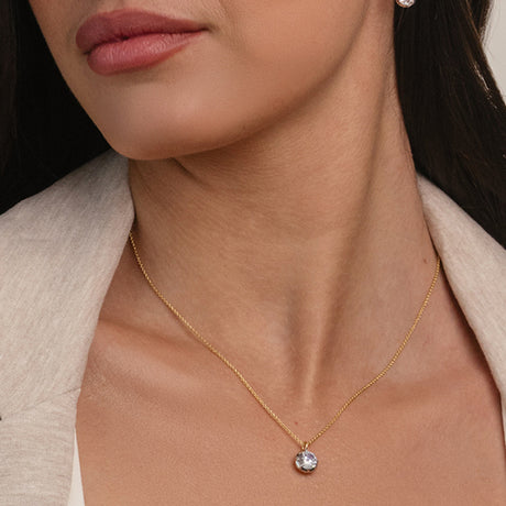 A close-up of a woman wearing a delicate gold chain necklace with a small round clear gemstone pendant, styled with a light beige blazer.