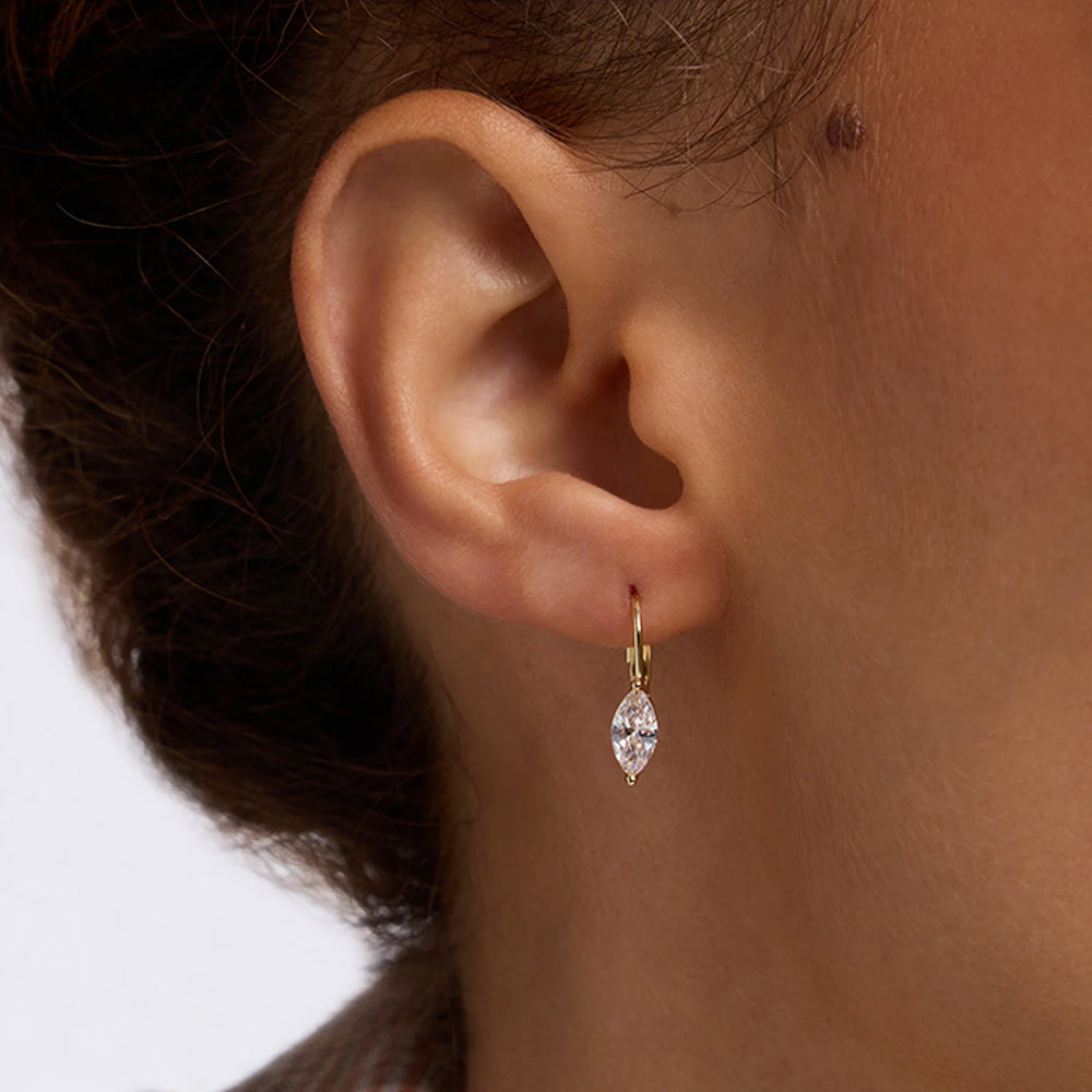 A close-up of a person's ear wearing a small gold hoop earring with a marquise-cut clear gemstone dangling below.