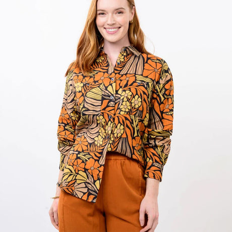 A woman wearing a long-sleeve button-up shirt with a vibrant orange, yellow, and black botanical pattern, paired with rust-colored pants, against a plain white background.