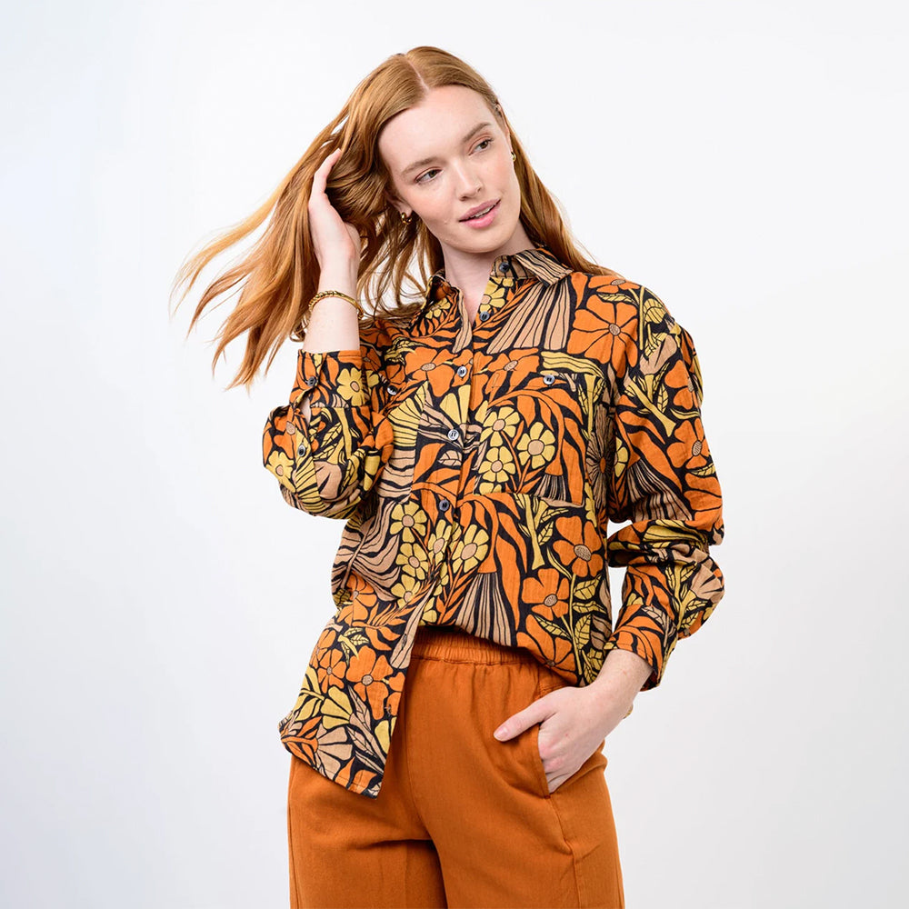 A woman modeling a long-sleeve button-up shirt with a bold retro floral and abstract pattern in orange, yellow, black, and brown, paired with rust-orange pants, against a plain light gray studio background.