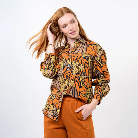 A woman modeling a long-sleeve button-up shirt with a bold retro floral and abstract pattern in orange, yellow, black, and brown, paired with rust-orange pants, against a plain light gray studio background.