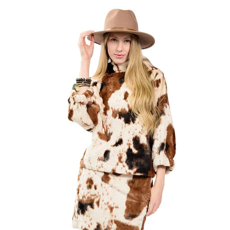 A woman modeling a faux fur two-piece outfit with a brown and cream cow print pattern, paired with a tan wide-brim hat, against a solid white background.