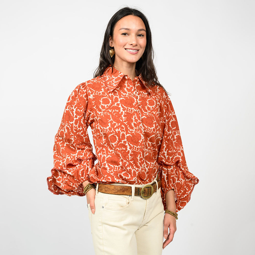 Woman wearing a patterned orange blouse and white pants on a plain background