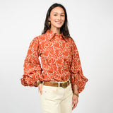 Woman wearing a patterned orange blouse and white pants on a plain background