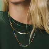 A close-up of a person wearing a layered necklace set. The set includes a gold paperclip chain with green links, a thin gold band, and a longer gold chain decorated with colorful beads and charms.