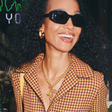 A close-up lifestyle shot of a smiling woman with curly hair wearing black sunglasses, a brown and red houndstooth blazer, and gold jewelry.