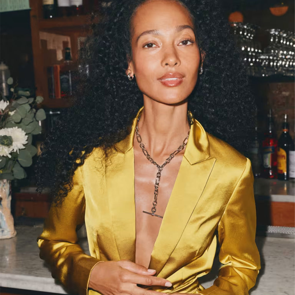 A woman with voluminous curly black hair wearing a shiny gold satin blazer and a gold chain necklace, photographed in a warm, dimly lit indoor setting with a bar or cafe background.