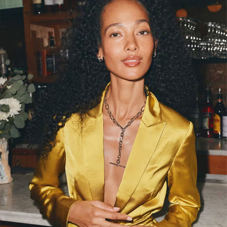 A woman with voluminous curly black hair wearing a shiny gold satin blazer and a gold chain necklace, photographed in a warm, dimly lit indoor setting with a bar or cafe background.