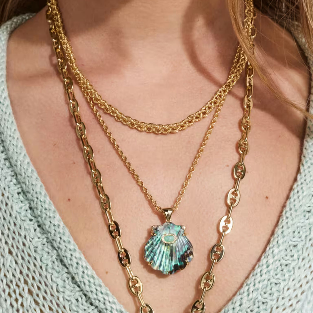 A close-up of a person wearing a layered necklace set featuring a scallop shell pendant with iridescent blue-green abalone inlay, layered over gold chains on a light blue knit top.