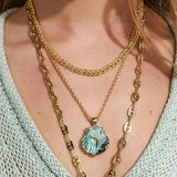 A close-up of a person wearing a layered necklace set featuring a scallop shell pendant with iridescent blue-green abalone inlay, layered over gold chains on a light blue knit top.