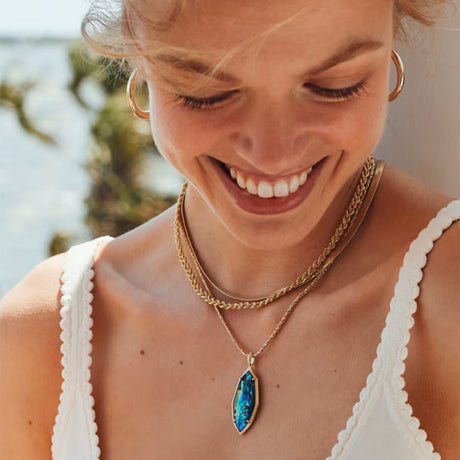 A close-up lifestyle shot of a smiling woman wearing a gold layered necklace with a marquise-shaped iridescent blue pendant, along with gold hoop earrings.