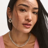 Close-up of a woman wearing a silver double-hoop earring, layered silver chain necklaces, and a pink top against a clean light background.