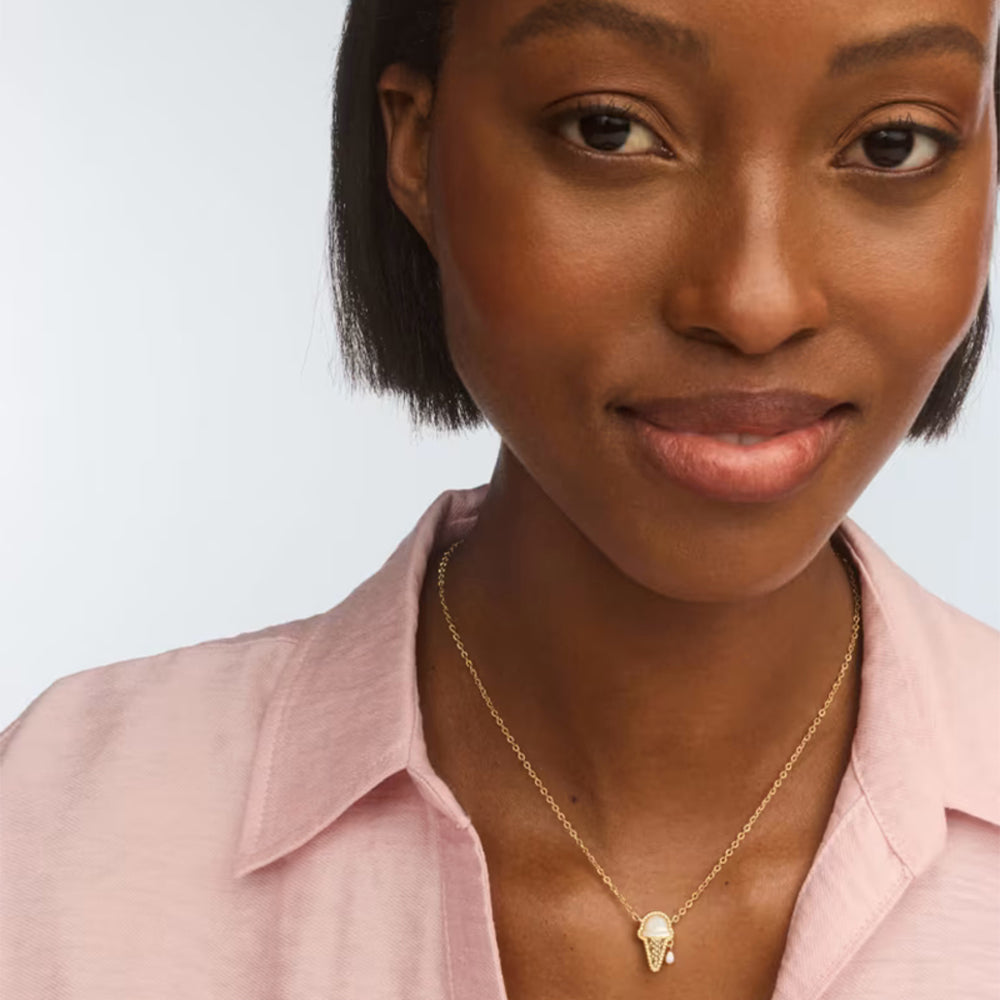A close-up of a woman wearing a delicate gold chain necklace with a small gold pendant, paired with a light pink collared shirt against a light gray background.