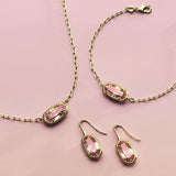 A jewelry set featuring a gold-tone chain necklace, bracelet, and drop earrings, each adorned with a pink marquise-cut gemstone in a gold-tone bezel setting, arranged on a solid pink background.
