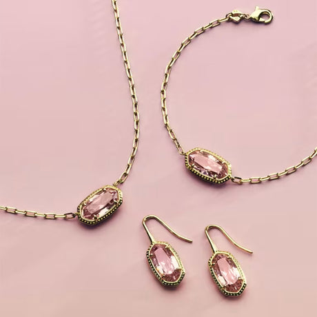 A jewelry set featuring a gold-tone chain necklace, bracelet, and drop earrings, each adorned with a pink marquise-cut gemstone in a gold-tone bezel setting, arranged on a solid pink background.