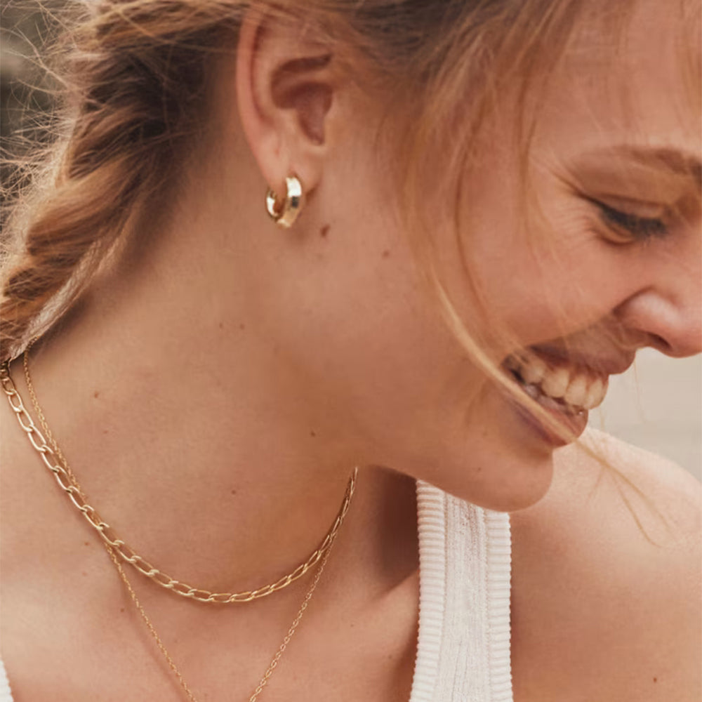 A close-up lifestyle shot of a woman wearing a small gold huggie hoop earring, layered with delicate gold necklaces, wearing a white ribbed tank top.