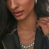 Close-up of a model wearing layered silver chain necklaces, including a delicate cable chain, a paperclip chain, and a chunky link chain, styled with a statement silver bib necklace and silver rings.