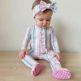 A baby sitting on a light wood floor wearing a pastel rainbow chevron-patterned footed sleeper and a matching bow headband.