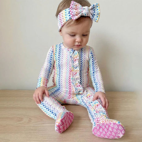 A baby sitting on a light wood floor wearing a pastel rainbow chevron-patterned footed sleeper and a matching bow headband.
