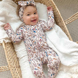 Baby in a floral onesie and headband lying in a crib with white bedding.