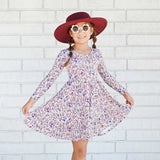 Young girl in a floral dress and red hat posing against a white brick wall.