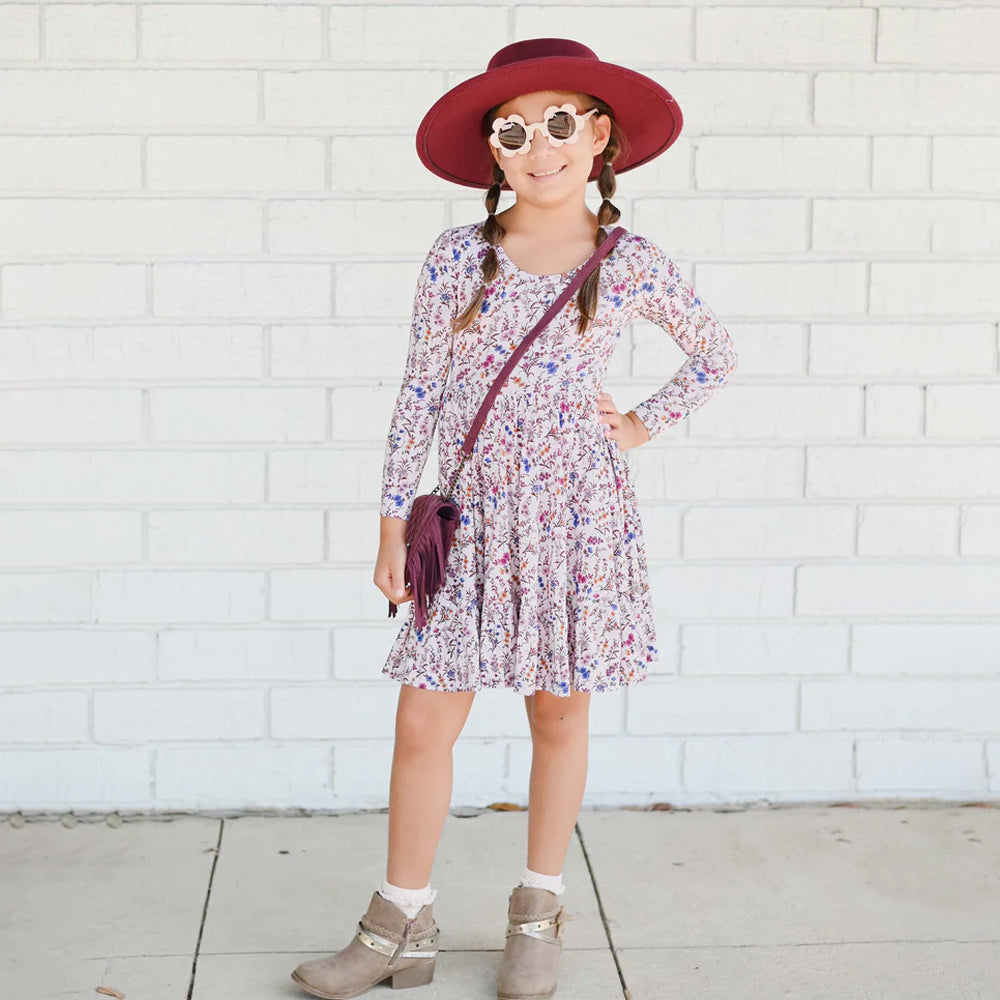 Young girl in a floral dress, red hat, and sunglasses standing against a white brick wall.